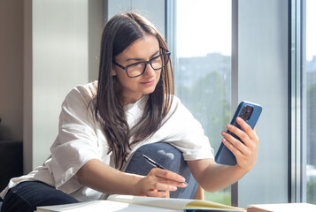 Student woman with smartphone and notepads at the window in the morning.