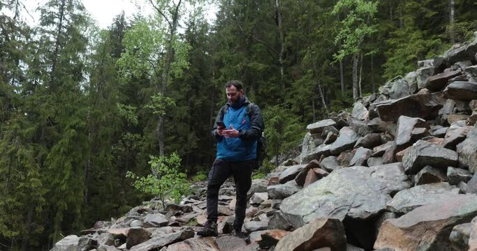 A man stands in the forest on large stones and takes out a smartphone to search for a network and make a call. Hiking in the mountains in rainy weather. He has a backpack on his shoulders.