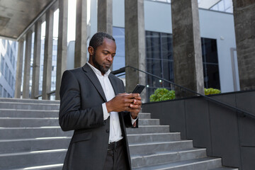 Serious thinking businessman holding phone outside office building, african american man reading focused message and checking news in app online.