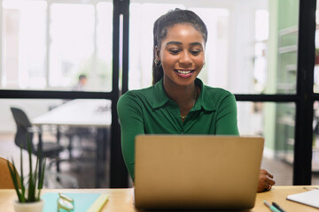 Friendly and positive african-american female employee or student using laptop, multiracial business woman looking at the screen, typing, responding to emails sitting in contemporary office