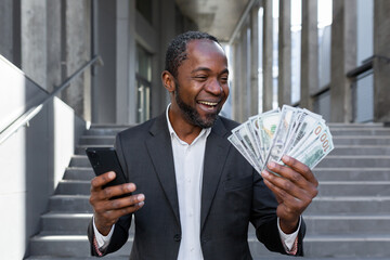 Mature African American businessman is happy about the successful results of financial activity, man is holding in his hands a smartphone and cash American money dollars, outside the office building.