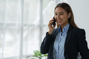 Happy Asian businesswoman talking on the phone and smiling while sitting at work using smartphone to talk. Contact via online application, concept of management