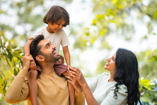 Happy indian parents playing with daughter while kid on father shoulder at outdoor - concept of parenthood, family bonding and weekend holidays
