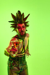 Fototapeta premium Young shirtless man, pun in extraordinary hairstyle drinking cocktail, posing against green studio background in neon light. Cheers. Concept of music, lifestyle, subculture, art, youth, human emotions