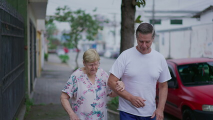 Adult son helping elderly mother to walk on sidewalk. Man caring and supporting senior person. Old age exercise routine, daily walk