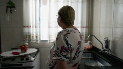 Back of contemplative elderly woman standing at kitchen by window. Pensive thoughtful real life old...