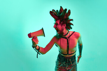 Young expressive shirtless man, punk souting in megaphone with agression against blue studio...