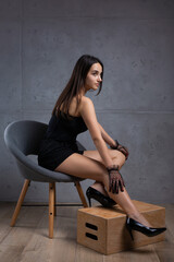 Fototapeta na wymiar Classic dark studio portrait of a young brunette woman in black clothes who is sitting on a chair against a concrete wall.