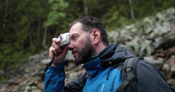 A man, close-up portrait, takes a photo of a landscape with a small camera in the mountains in rainy weather.