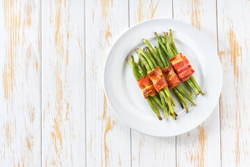 String beans wrapped in a roasted slices of bacon in a ceramic plate on a white wooden table, top view.