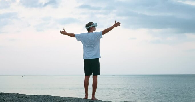 Young man in virtual reality glasses at sunset, raises his hands in the air, spins, rejoicing at the sunset on the sea, in augmented reality using Virtual Reality Headset. Virtual summer vacation