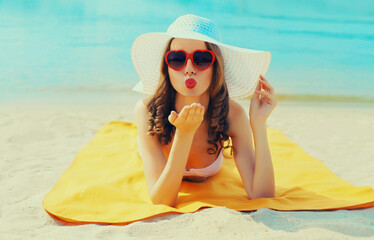 Summer vacation, happy relaxing young woman blowing her lips sends kiss in red heart shaped sunglasses, straw hat lying on sand on the beach on sea background
