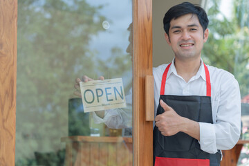 Business owner thumbs up to show his readiness to welcome customers. waitress man turning on open sign board on glass door in modern cafe coffee shop. New normal, small business, cafe, life style.