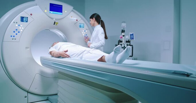 Medical worker adjusts capsule of MRI examination. Patient lies on MRI table and undergoes magnetic resonance imaging. Man in robe and protective cap enters capsule for examination.