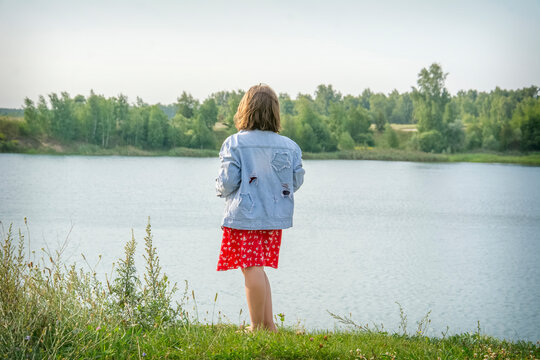 In summer, a girl in a denim jacket and dress stands on the river bank.