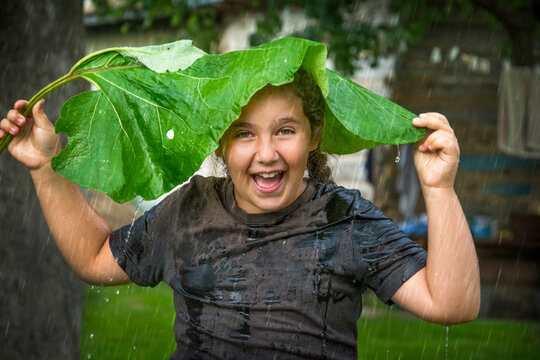 On a rainy summer day, a little girl hides from the rain under a large burdock leaf.