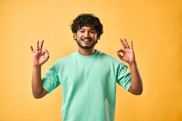 Handsome Indian man showing ok gesture, looking at camera and smiling while standing against yellow...