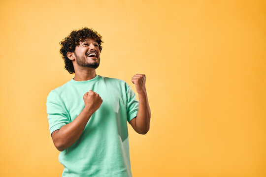 Emotion Of Joy, Celebration Of Victory. Handsome Indian Man Is Sincerely Happy, Looking To The Side And Making A Winning Gesture While Standing Against A Yellow Background. Copy Space.