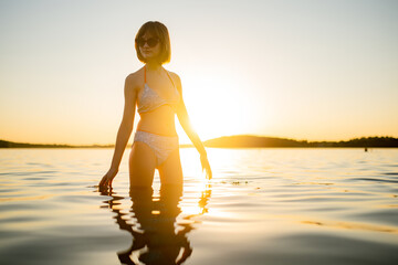 Fototapeta premium Silhouette of beautiful teenage girl having fun by a lake on warm and sunny summer evening. Pretty young girl on a sunset.