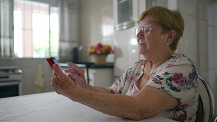Senior lady scrolling through social media using phone at home kitchen, domestic lifestyle of an...