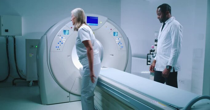 African-American doctor invites female patient to examination table. Female patient goes to MRI capsule. Female undergoes magnetic resonance imaging scan. Multicultural doctor performs examination.