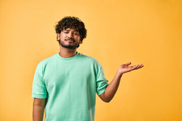 Emotion of choice, doubt. A handsome Indian man stands on a yellow background and points to space with one hand, grimacing at the camera. Copy space. - obrazy, fototapety, plakaty