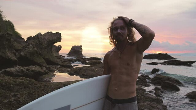 Wet sexy blonde long-haired surfer after surfing session on seashore at sunset waves his wet hair. Causacian man at sunset after surfing session waves his wet hair. End of surfing session at sunset.