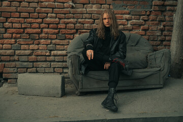 Young male model brutal man wearing black clothes sitting on old sofa on city street