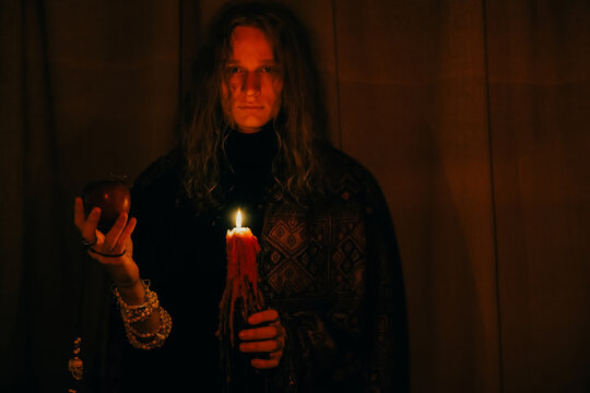 Portrait of young man with long blond curly hair holding a candle and an apple in his hands in a dark room. Loneliness, creativity, thoughtfulness