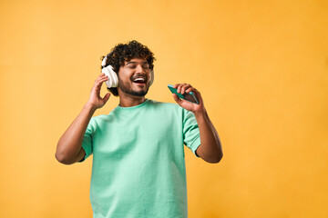 Portrait of a curly Indian man in a turquoise t-shirt singing with a phone in wireless headphones...