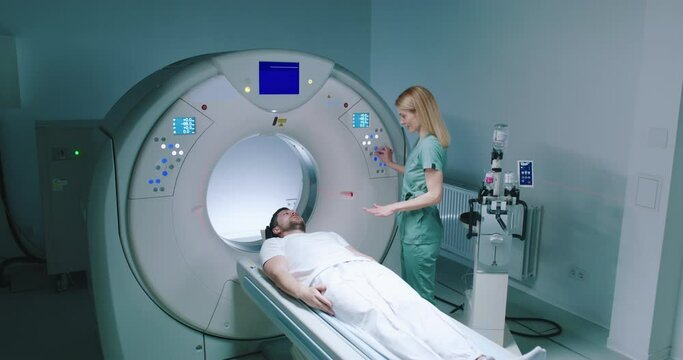 Male patient lies at TC scanner bed. Man waits for examine with MRI capsule. Female doctor dressed up in uniform explained to patient what to do. Medical worker perform magnetic resonance imaging.