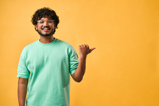 Portrait of a handsome Indian man with curly hair in a turquoise t-shirt and glasses standing on a yellow background and pointing to the side. Copy space.