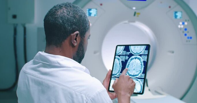 African American doctor examine scan attentively. Medical worker shooted from back. Male doctor examines MRI scan. Multicultural doctor is scrolling screen of tablet with scans of brain.