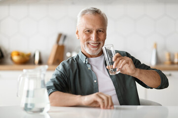 Senior Gentleman Holding Glass With Water While Sitting At Table In Kitchen