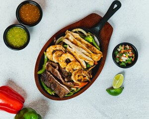 Top view of mixed meat fajitas in a pan on a wooden board with salsa, pico de gallo, and peppers