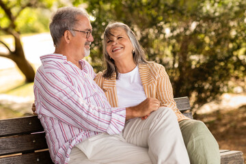 Cheerful glad european mature couple sit on bench, enjoy date together in park, talk at weekend in...