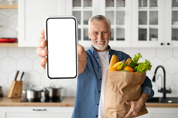 Grocery Delivery App. Smiling Senior Man Showing Blank Smartphone And Holding Groceries