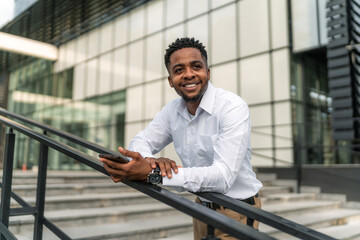 A portrait of a handsome Black businessman leaning against a railing during his break, holding a phone in his hand. With a relaxed posture and a thoughtful expression.