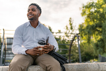 Obraz premium After a hard day at work, a young businessman, elegantly dressed, sits on the promenade, holding a cup of coffee and a mobile phone. With a relaxed demeanor, he takes a moment to relax.