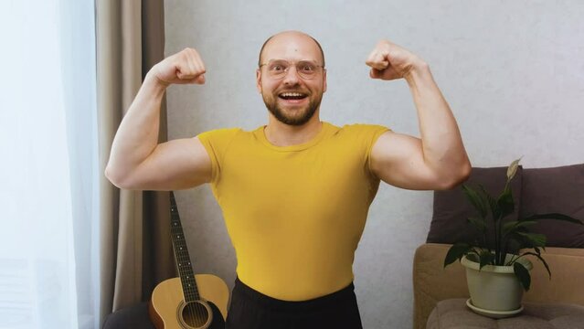 Funny athletic nerdy man with glasses looks at the camera, smiles stupidly and poses, showing muscles