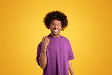 Fototapeta na wymiar Cheerful black adult curly man in purple t-shirt makes victory gesture, celebrates success