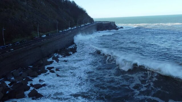 View of rough crashing waves rocks under a road with cars uder forest trees