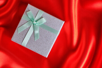 Silver gift box with a green ribbon and bow on a red fabric, silk background.