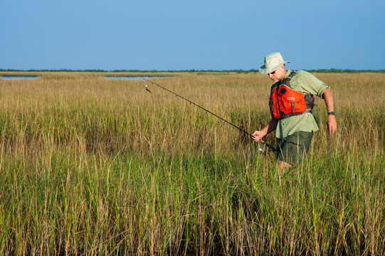 An older man with a fishing rod wades and wearing a red life jacket through marsh grass in Bayou Thunder Von Tranc, Louisiana
