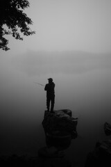 Greyscale shot of man standing on a rock in the middle of the lake