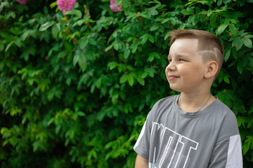 In summer, a child in a t-shirt boy stands against the background of green bushes. Copy space. Portrait of a child looking away with a wistful smile. Concept boy portrait on greenery background.