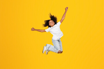 Cheerful curly teen black schoolgirl in white t-shirt has fun, jumping, freezes in air, enjoy...