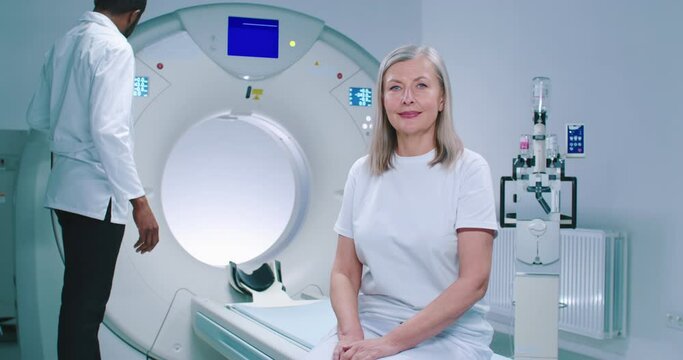 Shooting with smooth zoom. Mature woman sits at TC scanner bed. African American doctor adjusts MRI capsule behind woman. Woman dressed up in white is looking at camera.