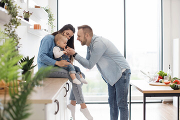 Charming mother on countertop hugging cute kid while smiling dad tickling baby girl on panoramic...