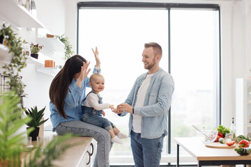 Happy parents keeping baby girl engaged while preparing food for dinner on sunny weekend. Attractive family man and wife miming hilarious stories for little daughter while staying in modern kitchen. - obrazy, fototapety, plakaty